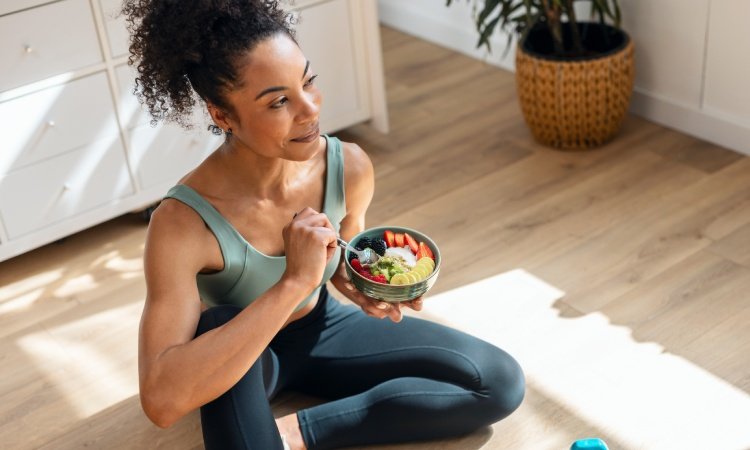 Sportliche Frau isst eine gesunde Obstschale, während sie zu Hause in der Küche auf dem Boden sitzt